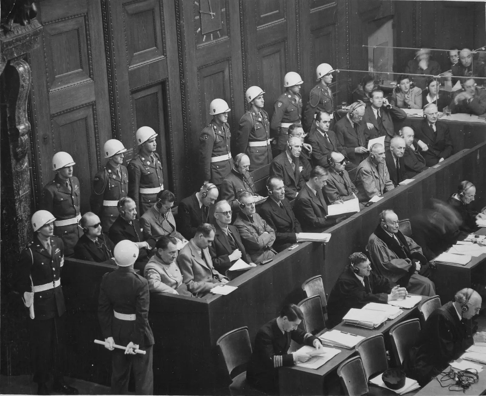 Black-and-white photo looking down on the dock in the Nuremberg courtroom, military police with white helmets
